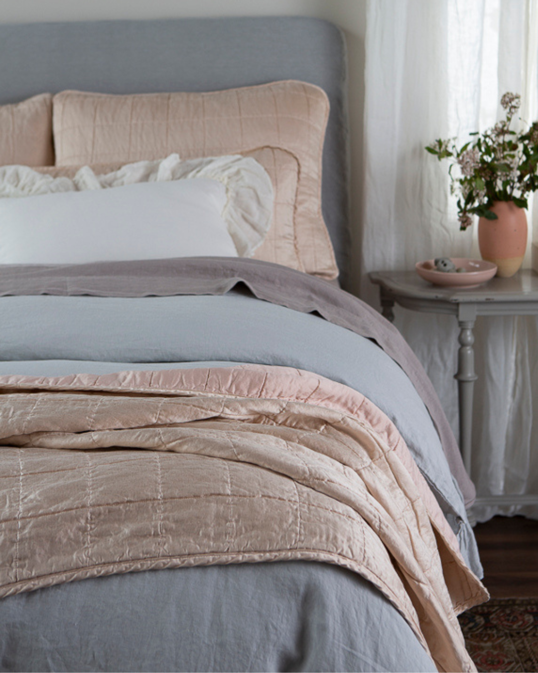 A soft palette and delicate ruffles unite with the structure of our square-quilt collection for a modern farmhouse look to best suit your serenity sanctuary. A gray table with decor of a similar palette sits next to the bed.