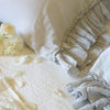 Linen Whisper Pillowcase (Single) | Linen Whisper pillow cases in cloud, with light cream toned sheets and flower petals - close-up overhead angle showcases ruffle detail trim.