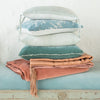 Ines Throw Pillow | Cloud | Ines 24 by 24 pillow stacked neatly with throw pillows and blankets in blue, pink, and green tones, against a plain background - side view.