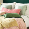 Taline throw pillows and blanket in green and gold tones shine against pink-toned embroidered linen - cropped three-quarter angle.