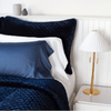 Silk velvet quilted shams in deep midnight blue with matching coverlet, layered simply with white and midnight sleeping pillows - cropped three-quarter angle.