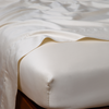 Bria Flat Sheet | Parchment | Cotton sateen flat sheet draped over matching fitted sheet. Shown from the top corner, the flat sheet is rumpled, highlighting the shine of the fabric.