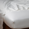 Bria Flat Sheet | Winter White | Cotton sateen flat sheet draped over matching fitted sheet. Shown from the top corner, the flat sheet is rumpled, highlighting the shine of the fabric.
