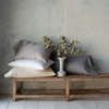 Bria Pillowcase (Single) | Bria sleeping pillows in various white and grey tones, stacked on a wooden bench against a textured neutral wall. A ceramic vase with greenery sits between the pillows.