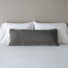 Carmen Throw Pillow | Fog | Silk velvet lumbar pillow with petite ruffle, leaning upright against neutral bedding and headboard.