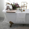 White Frida guest towel draped over the edge of an antique tub, with copper accents and lavender and green floral bouquet.
