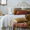Harlow coverlet in winter white on a neatly made bed against a grand tufted headboard. Warm brown and gold toned throw pillows and blankets in silk charmeuse and silk velvet provide contrast to the quilted cotton velvet - end of bed view.