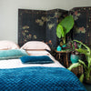 Linen shams and duvet cover in pearl, contrasted with silk velvet in blues and greens, dark floral wall panel, and tall green plants - foot of bed angle.