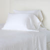Paloma Pillowcase (Single) | White | sleeping pillows leaning upright against a white wall on monochromatic sheeting - cropped three-quarter angle.