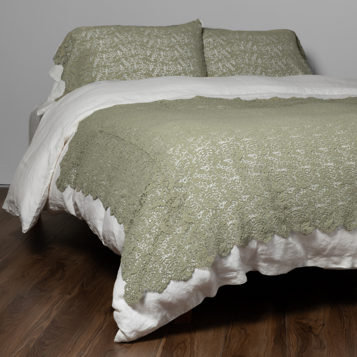 Allora Bed Scarf | Desert Sage | a lace throw blanket in desert sage, a serenely elegant green, offering a hushed hue, shown with matching pillowcases on a bed dressed in white and against a white background.