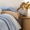 Linen Sham | a blue pillowcase lays atop a honey-colored sham on a bed made in light blue linen bedding with a decorative side table between the bed and window.