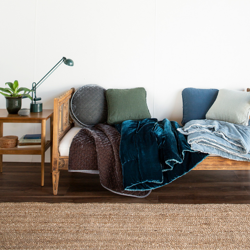 Austin Throw Pillow | A daybed bench against a white wall and dark wood flooring. A neutral rug ties in with the bench and side table. An assortment of throw pillows and blanket in grey and blue tones is the focus.