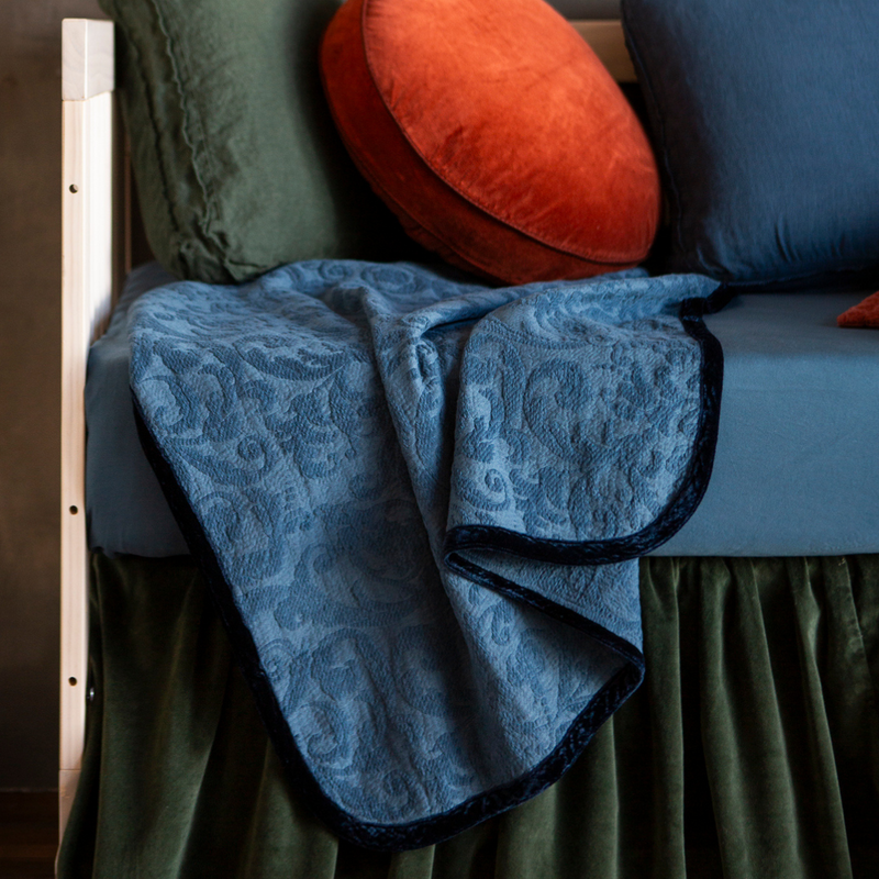 Adele Baby Blanket | Midnight | A close up of an Adele blanket in Midnight, a dark blue tone — shown with blue and green crib bedding and a rust colored round throw pillow.