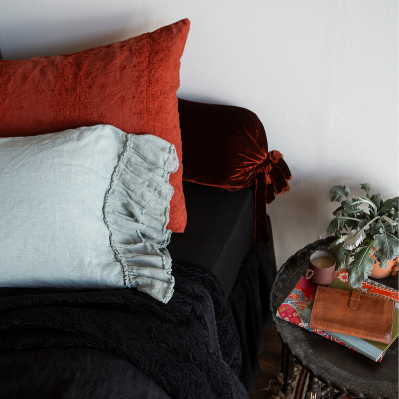 Linen Whisper Pillowcase (Single) | linen pillowcase with ruffle shown against an embroidered linen sham and silk velvet bolster pillow - shown with black sheets and coverlet against a white wall with side table.