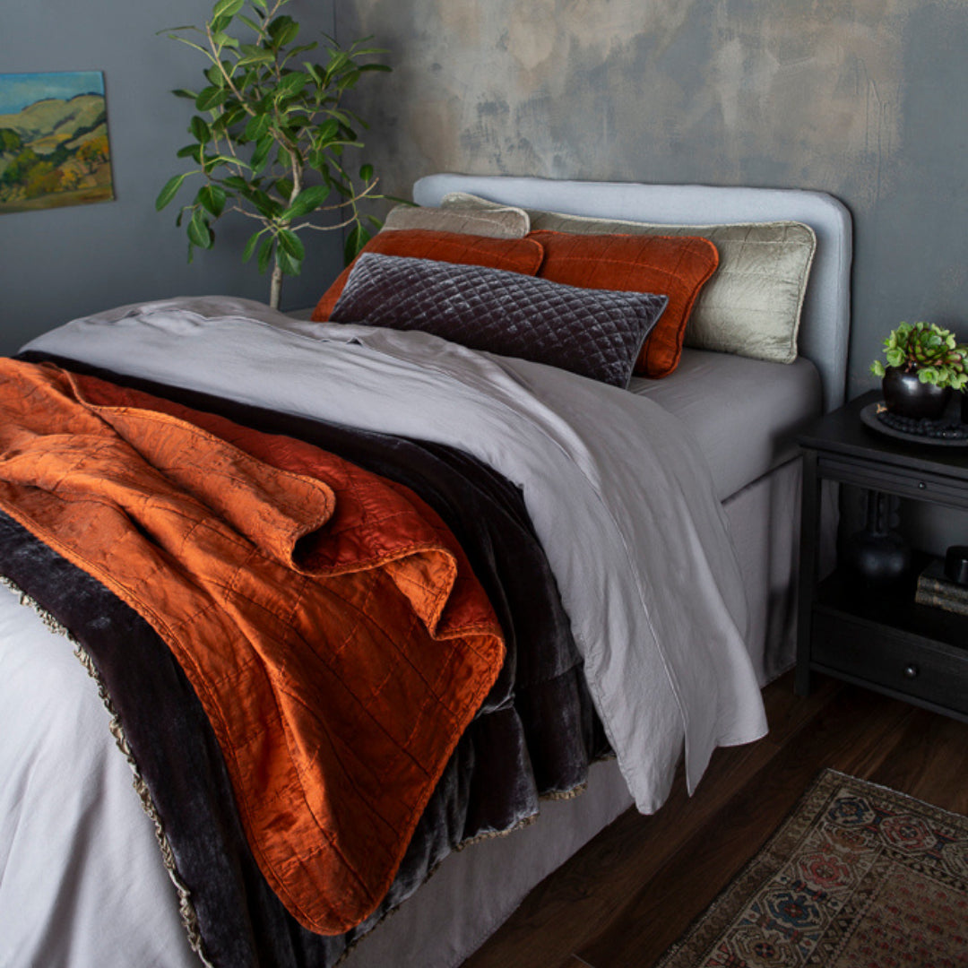 A bed dressed with Sel Throw Pillows in Moonlight (18x54 & 18x18) and Silk Velvet Quilted Throw Pillow (16x36) in Moonlight with Sel Shams in Autumn on a bed layered with Madera Luxe Sheets and Duvet Cover in Moonlight. Carmen Blanket at the bed end and Sel Throw Blanket in Autumn, finished with the Austin Bed Skirt in Moonlight. 