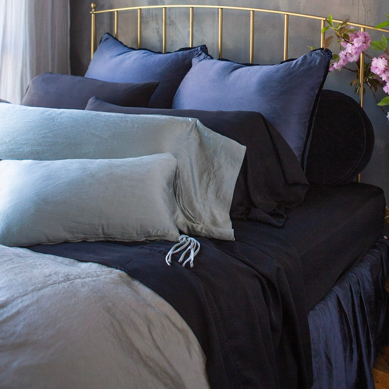 Paloma Sham | dark blue shams join a moody palette of black and blueish-green-grey accented by a gold headboard frame and light pink flowers.