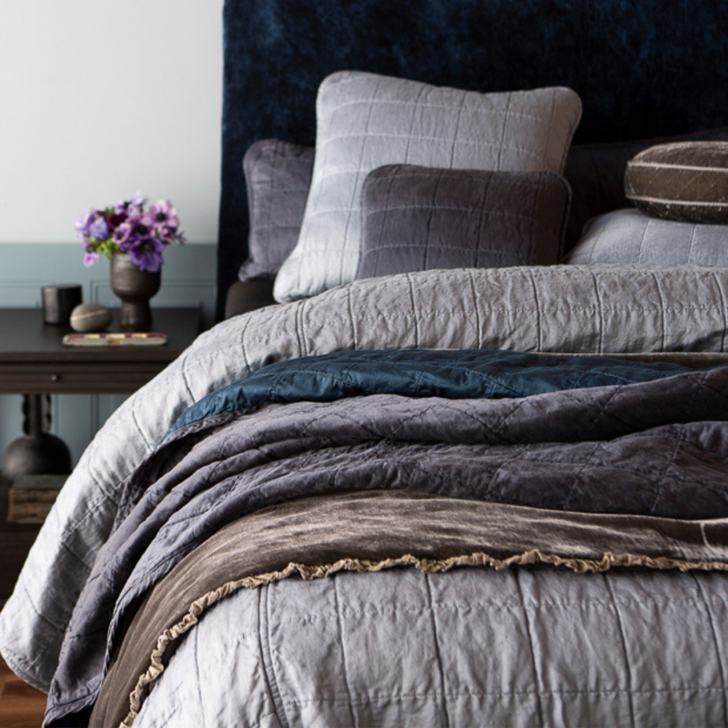 Sel Coverlet | a quilted charmeuse coverlet layered with a bed end blanket and throw blanket - shown from the foot of the bed, the pillows near the headboard are slightly out of focus as are the flowers on the side table.