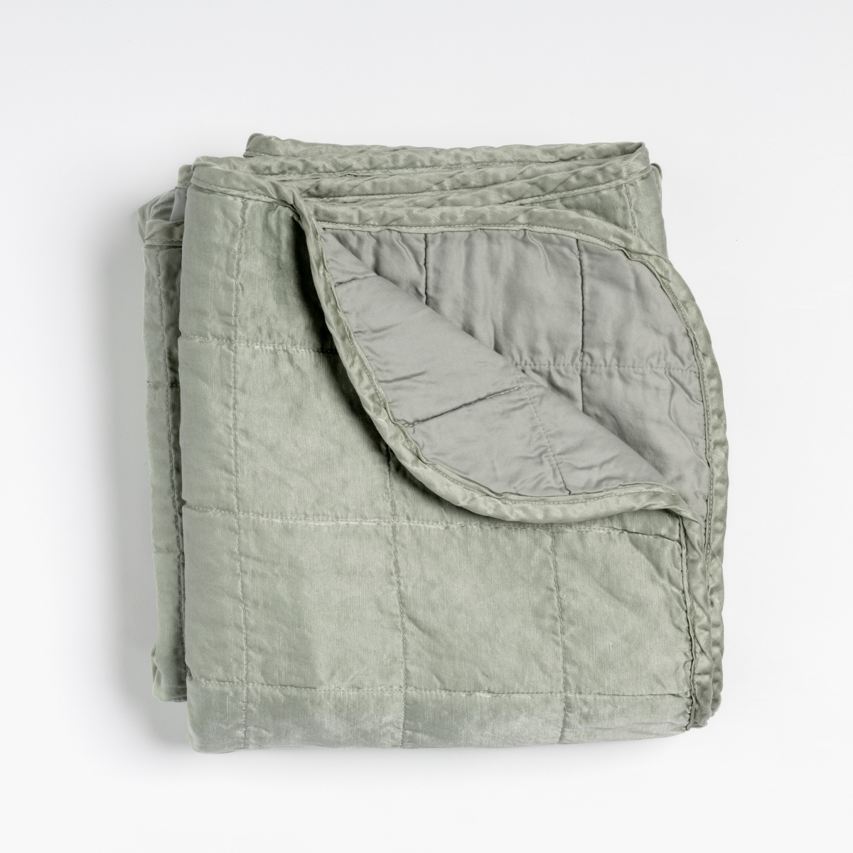 Sel Blanket | Eucalyptus | an overhead shot of a quilted charmeuse blanket in eucalyptus, a soft light green, with a corner folded down, showing its cotton sateen back.