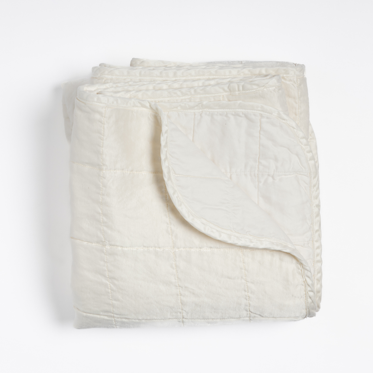 Sel Blanket | Winter White | an overhead shot of a quilted charmeuse blanket in winter white, a softer and warmer tone than our classic white, with a corner folded down, showing its cotton sateen back.