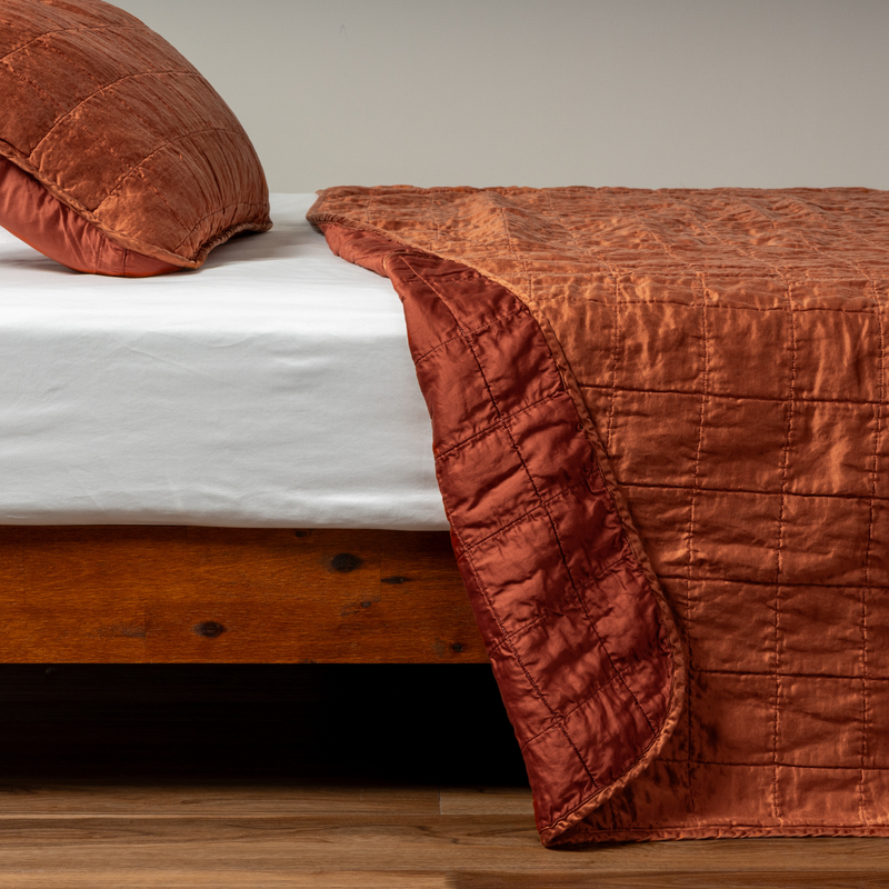 Sel Coverlet | Autumn | A side shot of a square quilted charmeuse coverlet in autumn, a rich terracotta tone, with its matching sham, the corner of the coverlet peeled back, revealing the cotton sateen back.