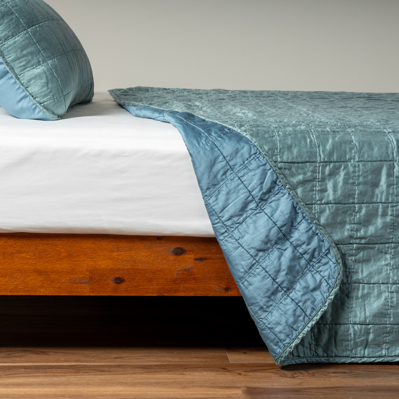 Sel Coverlet | Cenote | A side shot of a square quilted charmeuse coverlet in cenote, a vibrant ocean-inspired blue-green, with its matching sham, the corner of the coverlet peeled back, revealing the cotton sateen back.