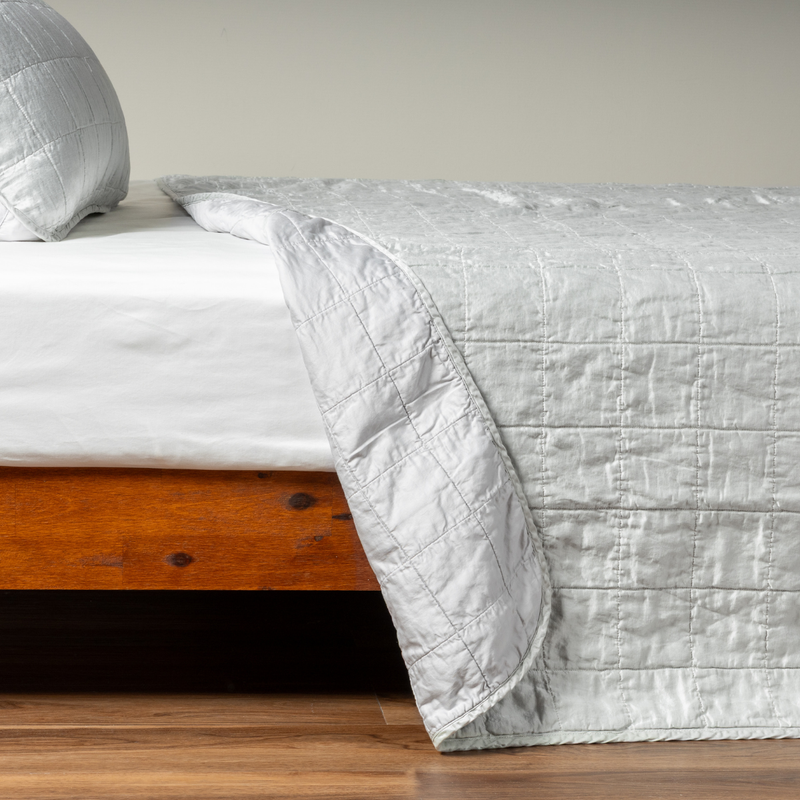Sel Coverlet | Cloud | A side shot of a square quilted charmeuse coverlet in cloud, a subtle sky blue-grey, with its matching sham, the corner of the coverlet peeled back, revealing the cotton sateen back.