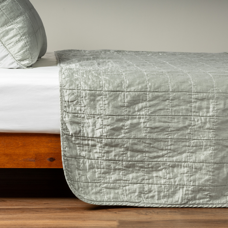 Sel Coverlet | Eucalyptus | A side shot of a square quilted charmeuse coverlet in eucalyptus, a soft light green, with its matching sham, the corner of the coverlet peeled back, revealing the cotton sateen back.