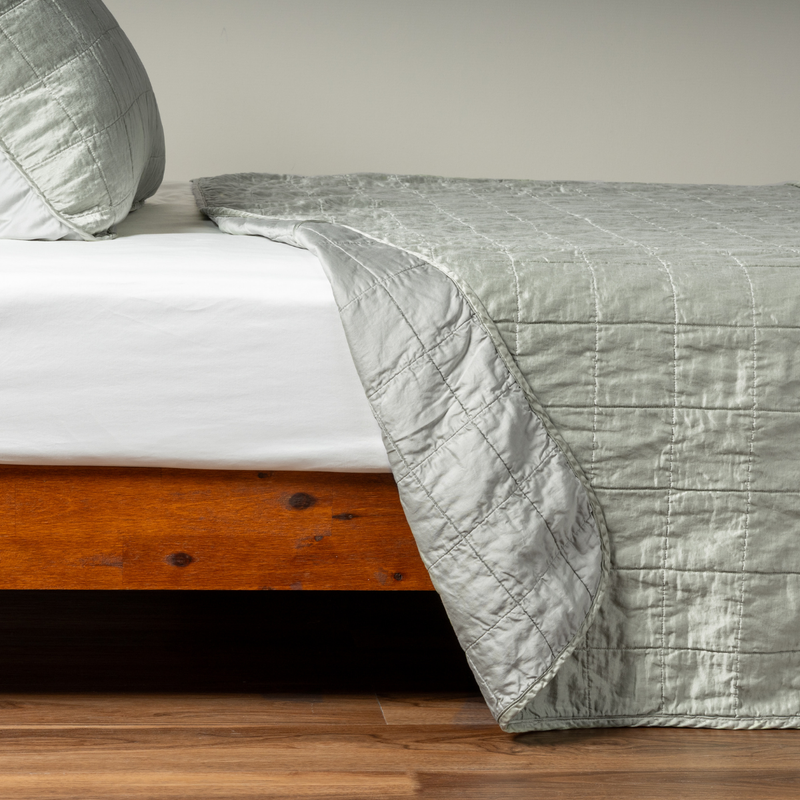 Sel Coverlet | Eucalyptus | A side shot of a square quilted charmeuse coverlet in eucalyptus, a soft light green, with its matching sham, the corner of the coverlet peeled back, revealing the cotton sateen back.