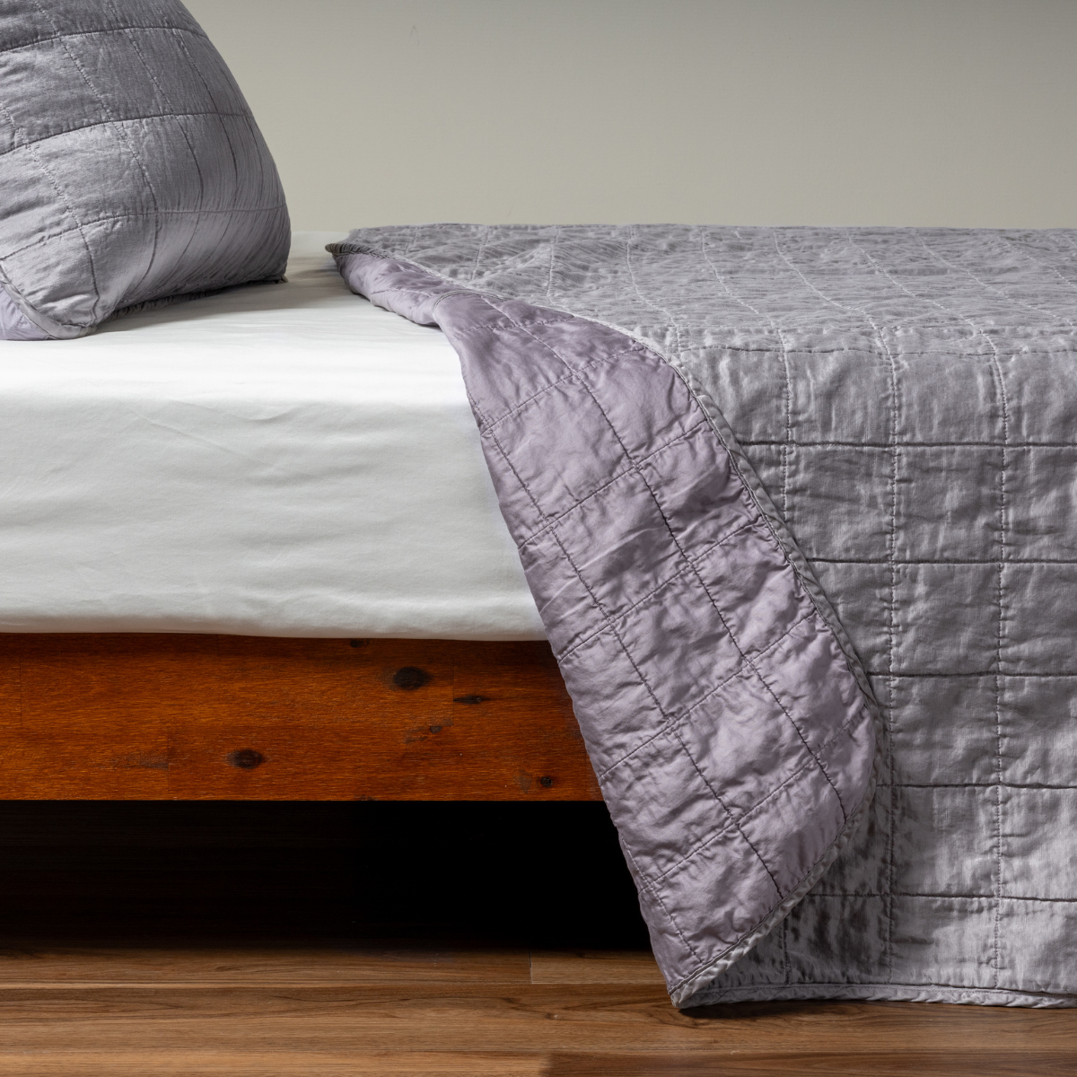 Sel Coverlet | French Lavender | A side shot of a square quilted charmeuse coverlet in french lavender, a neutral violet tone, with its matching sham, the corner of the coverlet peeled back, revealing the cotton sateen back.