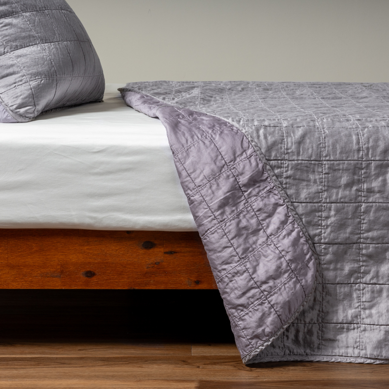 Sel Coverlet | French Lavender | A side shot of a square quilted charmeuse coverlet in french lavender, a neutral violet tone, with its matching sham, the corner of the coverlet peeled back, revealing the cotton sateen back.