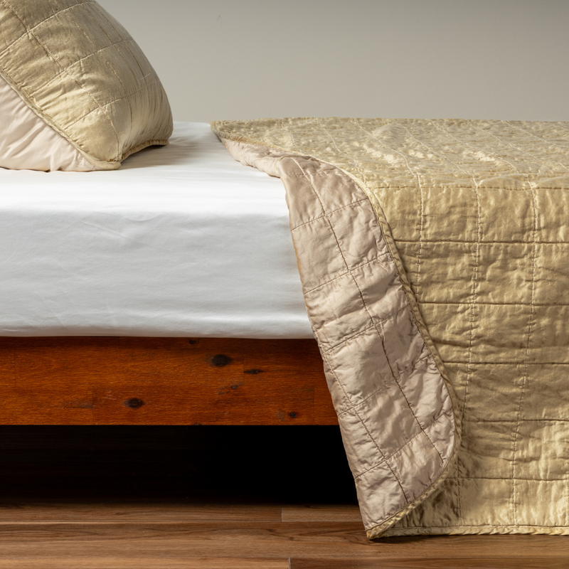 Sel Coverlet | Honeycomb | A side shot of a square quilted charmeuse coverlet in honeycomb, a warm golden tone, with its matching sham, the corner of the coverlet peeled back, revealing the cotton sateen back.
