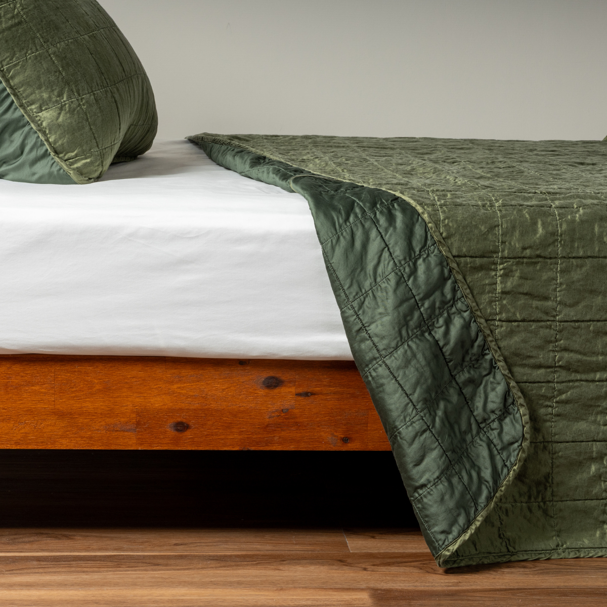 Sel Coverlet | Juniper | A side shot of a square quilted charmeuse coverlet in juniper, a deeply saturated dark green, with its matching sham, the corner of the coverlet peeled back, revealing the cotton sateen back.