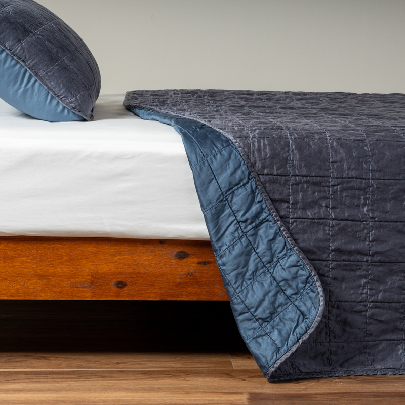 Sel Coverlet | Midnight | A side shot of a square quilted charmeuse coverlet in midnight, a rich indigo tone, with its matching sham, the corner of the coverlet peeled back, revealing the cotton sateen back.