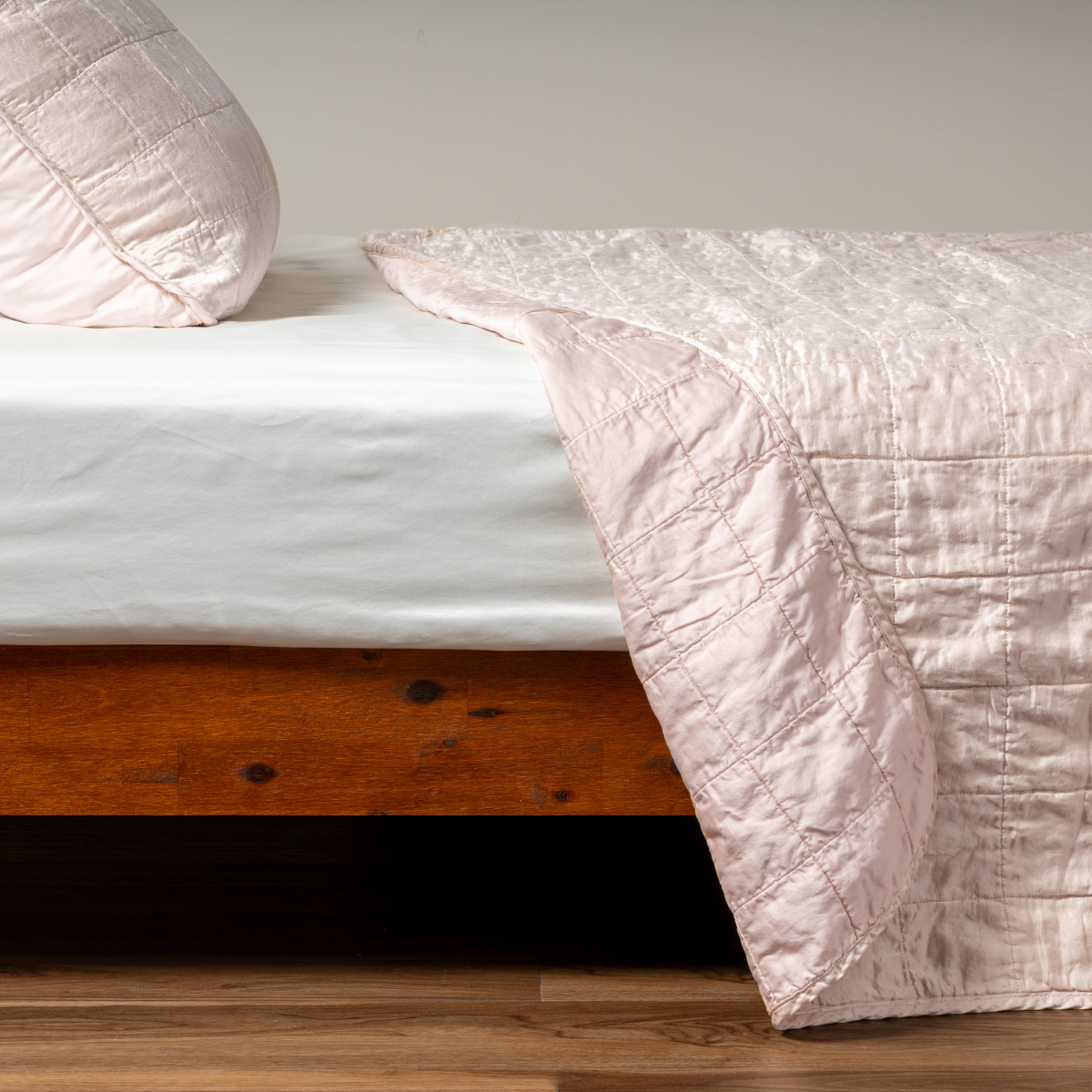 Sel Coverlet | Pearl | A side shot of a square quilted charmeuse coverlet in pearl, a soft rose pink tone, with its matching sham, the corner of the coverlet peeled back, revealing the cotton sateen back.