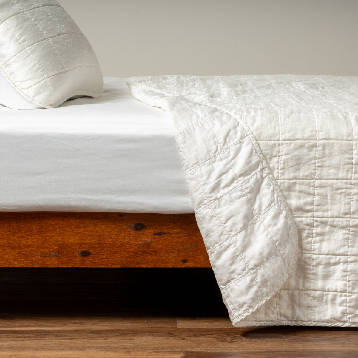 Sel Coverlet | Winter White | A side shot of a square quilted charmeuse coverlet in winter white, a softer and warmer tone than our classic white, with its matching sham, the corner of the coverlet peeled back, revealing the cotton sateen back.