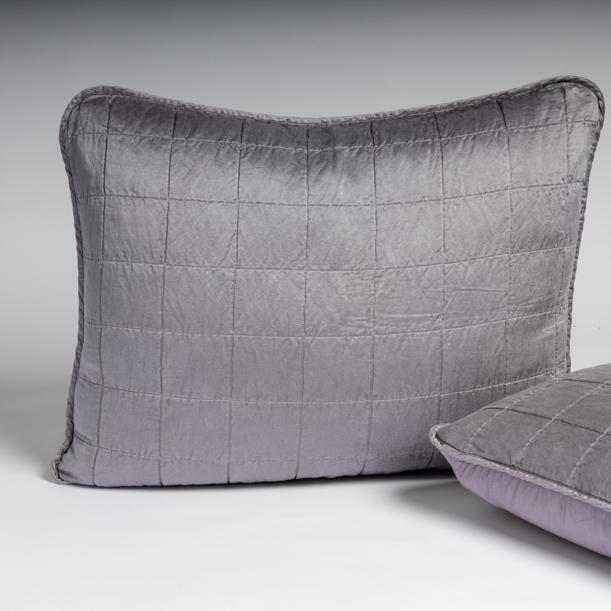 Sel Sham | French Lavender | A pair of shams shown in french lavender, a neutral violet tone, against a white background with one sham upright and the other on its side to show the cotton sateen back and square-quilted charmeuse front.