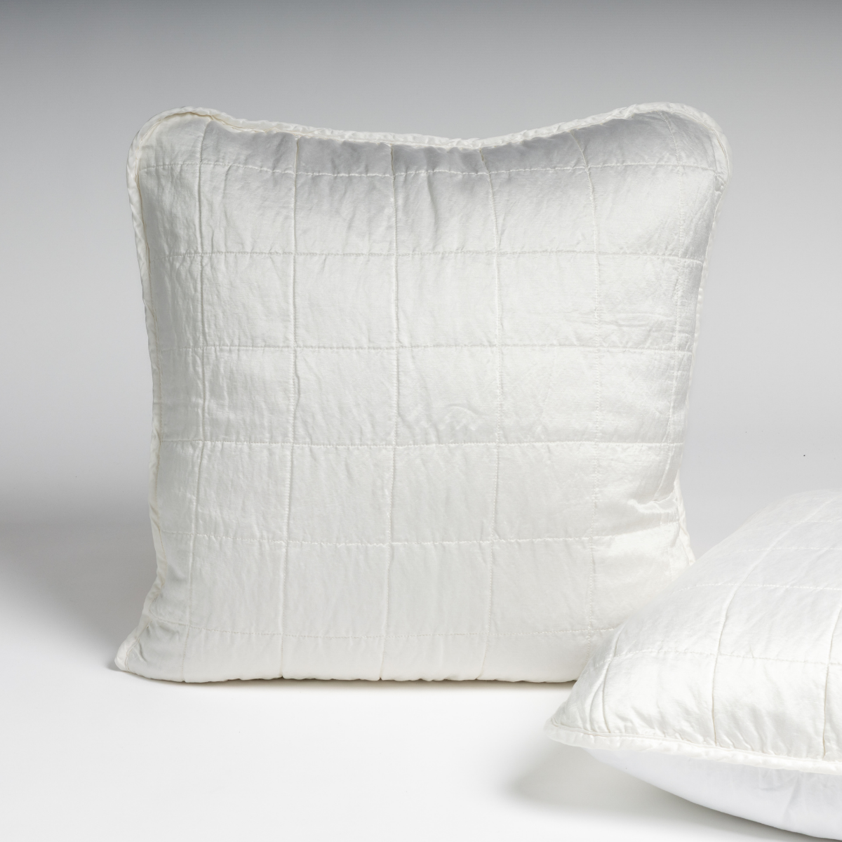Sel Sham | White | A pair of shams shown in white against a white background with one sham upright and the other on its side to show the cotton sateen back and square-quilted charmeuse front.