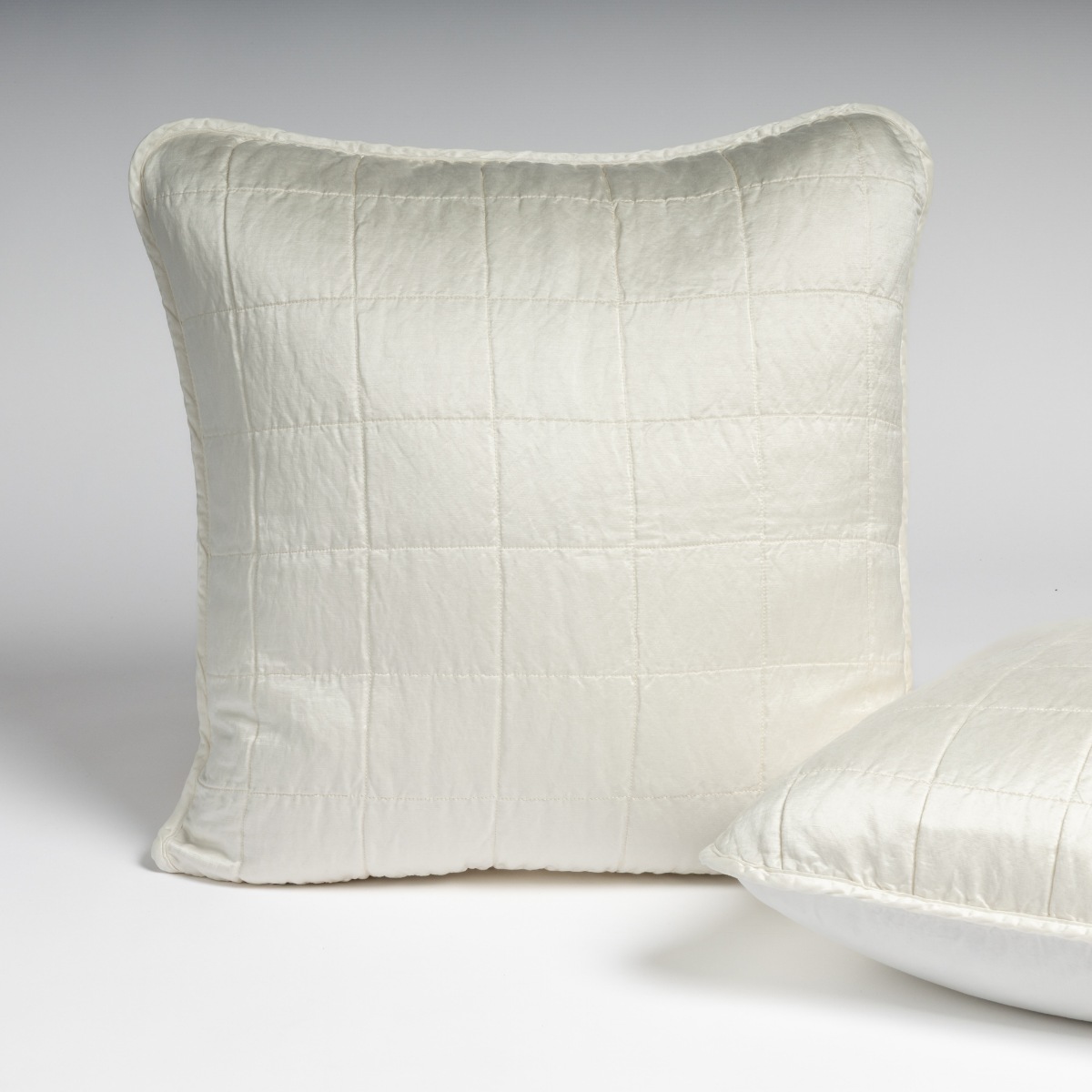 Sel Sham | Winter White | A pair of shams shown in winter white, a softer and warmer tone than our classic white, against a white background with one sham upright and the other on its side to show the cotton sateen back and square-quilted charmeuse front.