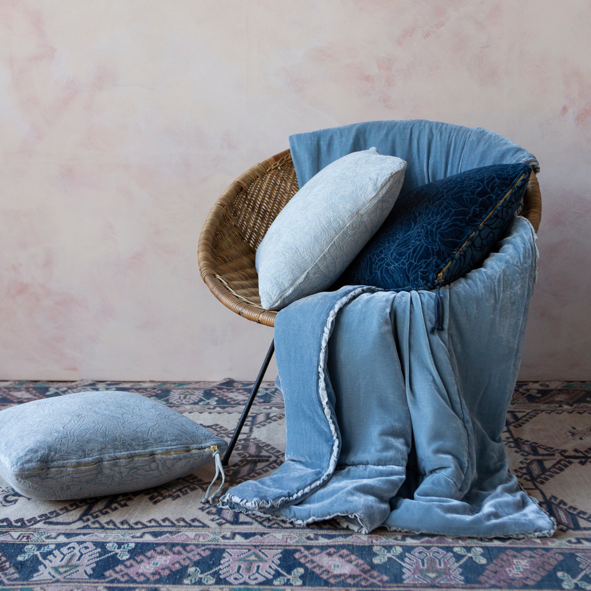 Georgia Throw Pillow | a papasan-shaped chair is draped with a silk velvet throw blanket and coordiinating chenille and cotton jacquard throw pillows in shades of blue — shown against a pink-toned wall and colorful rug.