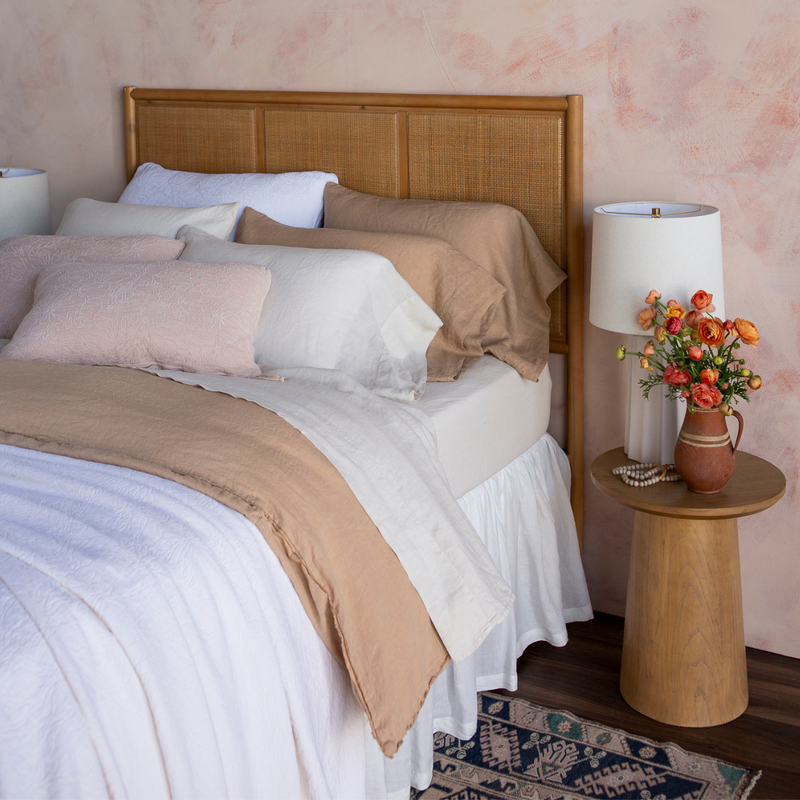 Georgia Bedspread | a three-quarter view of a fully dressed bed in light tones with a rattan headboard against a pink-toned wall.