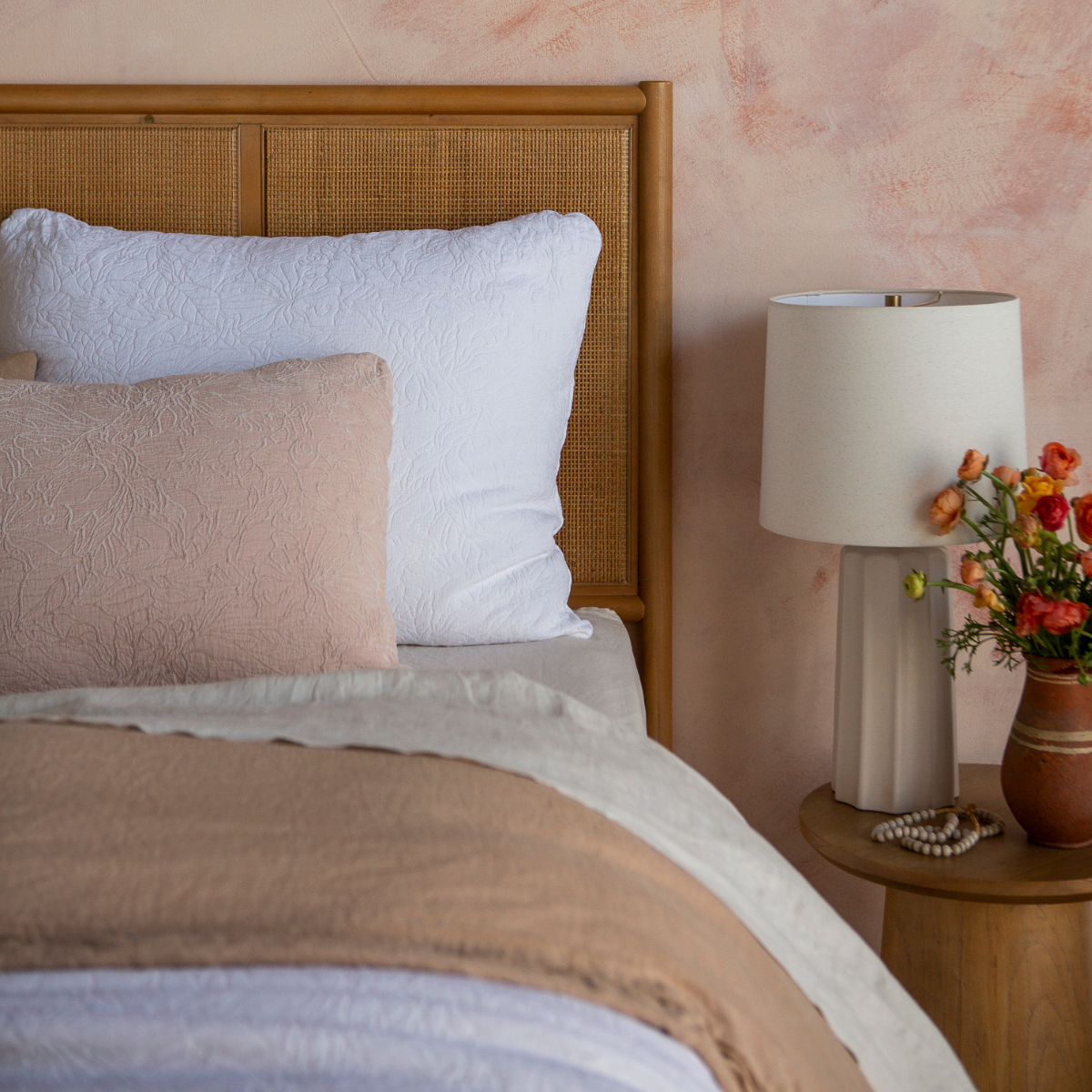 Georgia Sham | shot from the foot of the bed, the shams and throw pillows are shown against a rattan headboard with a pink-toned wall in the background. The side table has a neutral lamp and vase of colorful flowers