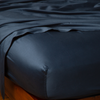 Bria Twin Flat Sheet | Midnight | Cotton sateen flat sheet draped over matching fitted sheet. Shown from the top corner, the flat sheet is rumpled, highlighting the shine of the fabric.