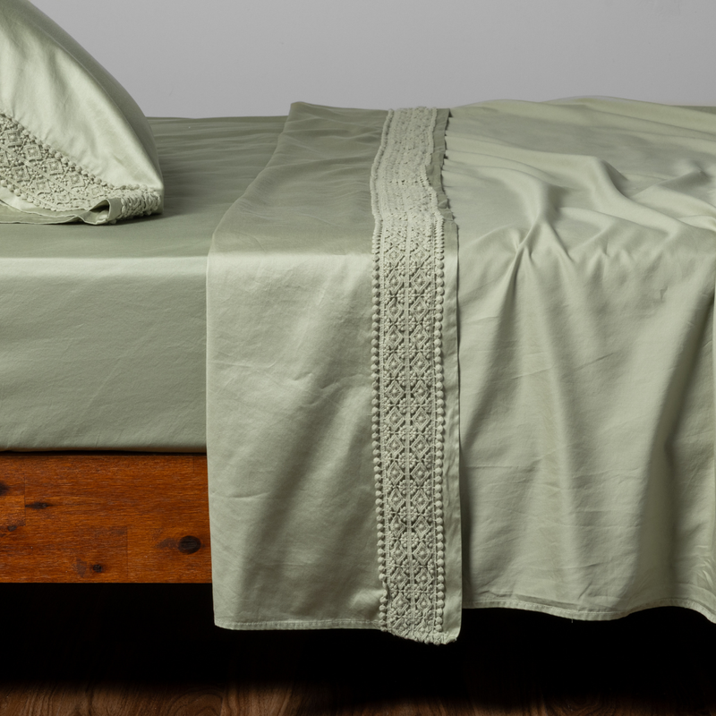 Bria Flat Sheet | Desert Sage | a matching flat sheet with lace, fitted sheet and  pillowcase in desert sage, a serenely elegant green, offering a hushed hue, shown from the side angle on a bed.