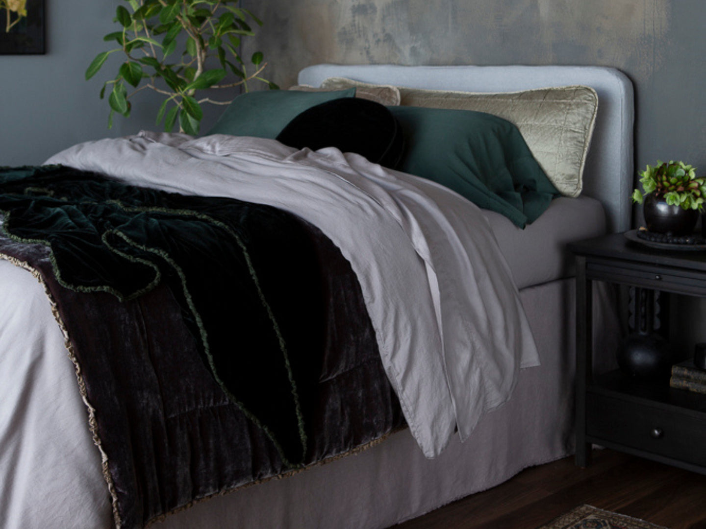 A moonlight inspired bed layered in the range of two main colors - moonlight and juniper. There is a moody backdrop for the bed with dark furniture in the foreground of the photo