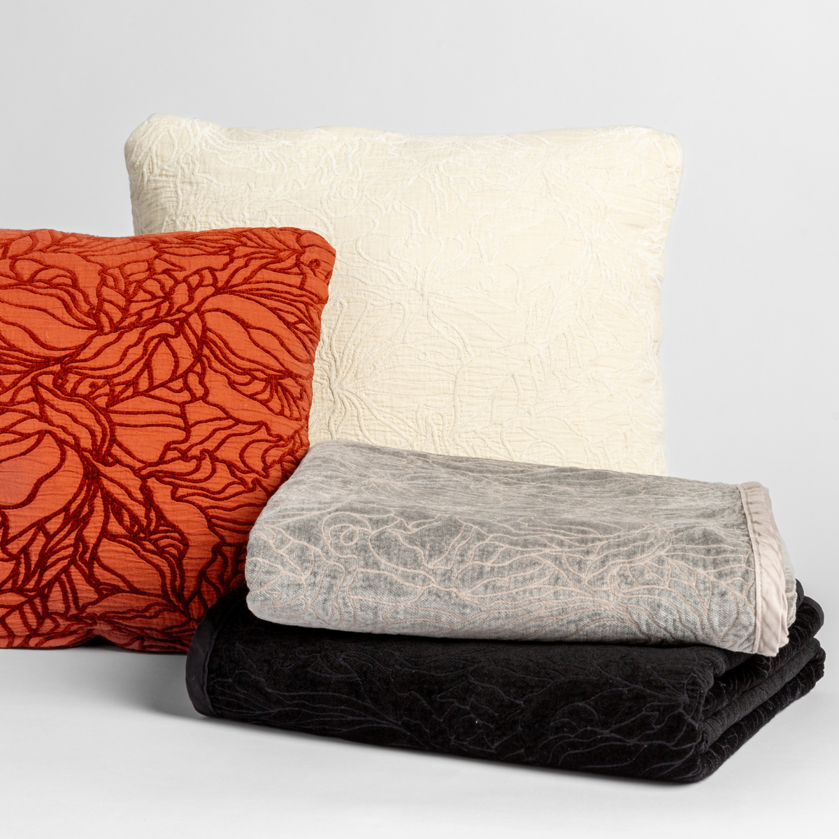 A cluster of two Georgia throw pillows displayed on their cotton jacquard side and two Georgia blankets displayed on their chenille jacquard sides, all against a white background, in the following colorways | one throw pillow in Autumn, a rich terracotta tone, another throw pillow in Winter White, a wooly and warm tone, one blanket in Fog, a neutral mid-grey, and another blanket in Corvino, a raven black tone.