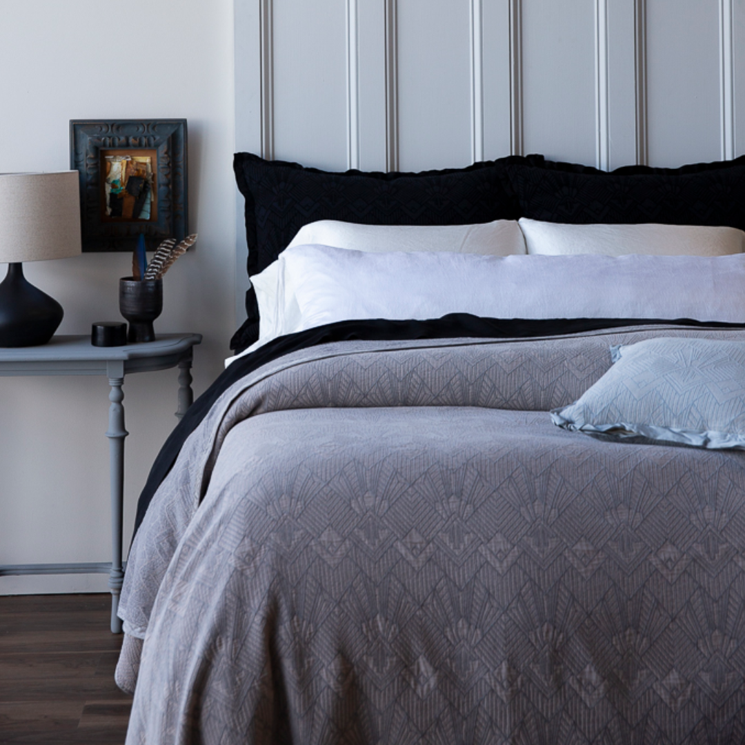 The Trettino Collection | Bedroom with a bed featuring gray bedding and black pillows, nightstand with lamp and decor.