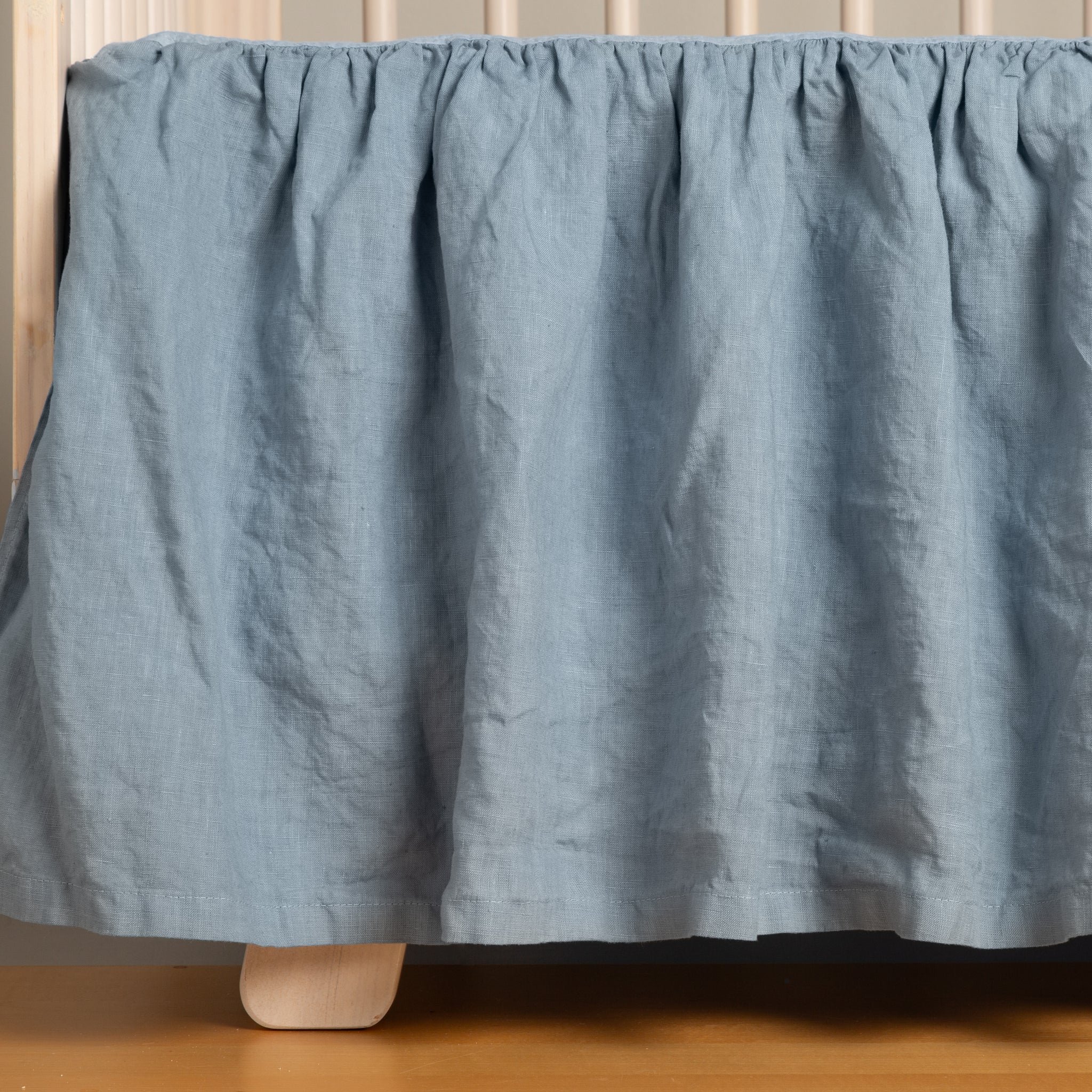 Linen Crib Skirt | Tahoe Blue | a crib skirt on a natural wood crib on a medium wood floor.
