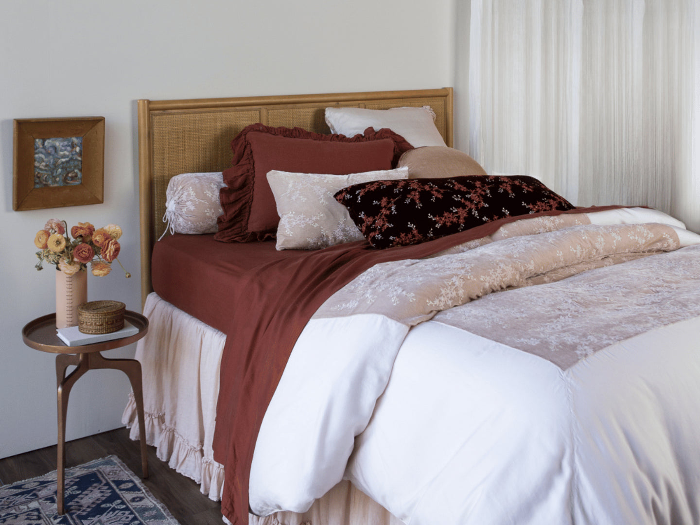 [allvariants]: three-quarter view of a dressed bed in rust and pale pink tones with a metallic side table holding a vase of flowers and trinket box. 