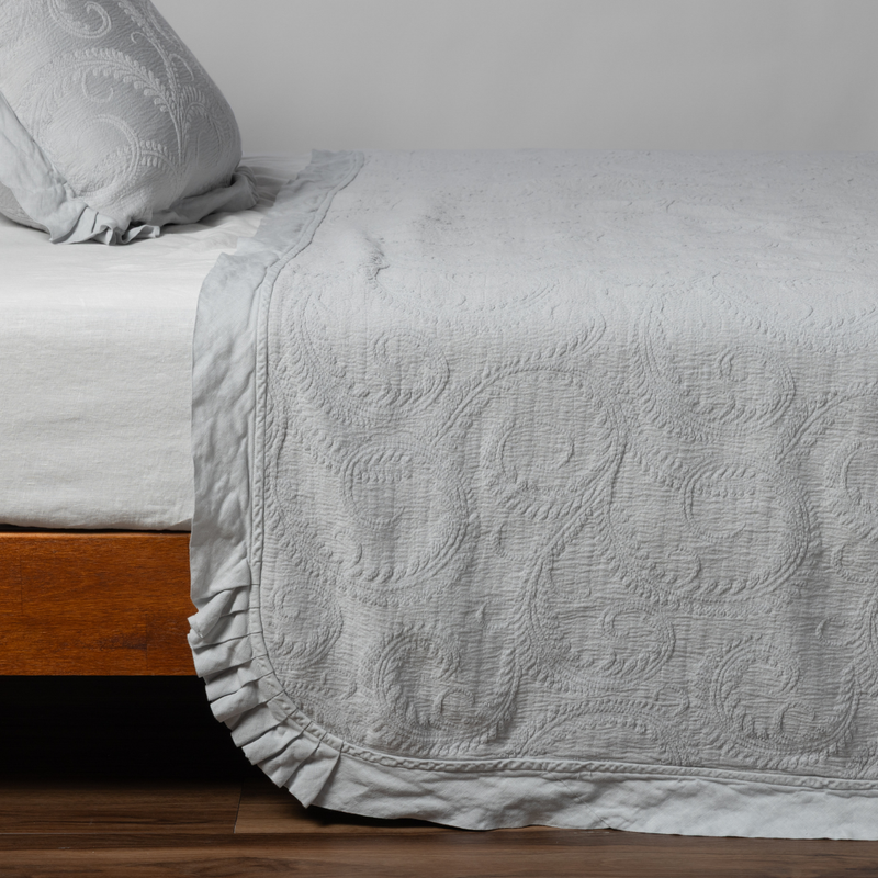 Marisette Bedspread | Cloud | matching organic cotton damask trimmed with linen bedspread and sham on a bed with white fitted sheet, shown from the side against a plain background.