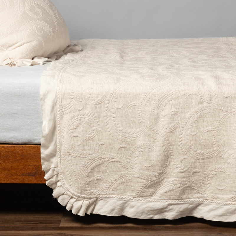 Marisette Bedspread | Natural | matching organic cotton damask trimmed with linen bedspread and sham on a bed with white fitted sheet, shown from the side against a plain background.